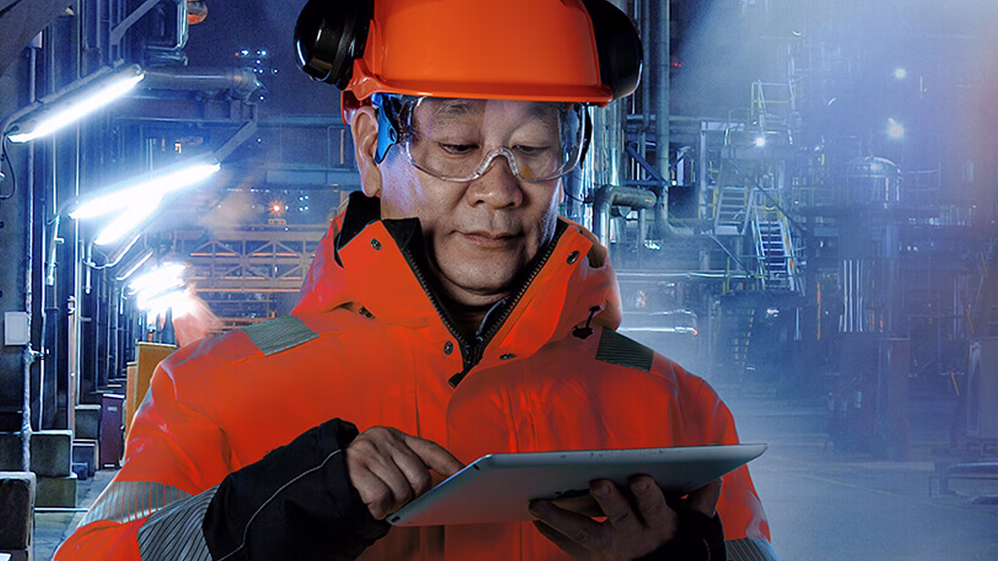 Werknemer met oranje helm en veiligheidsvest houdt tablet vast in industriële omgeving