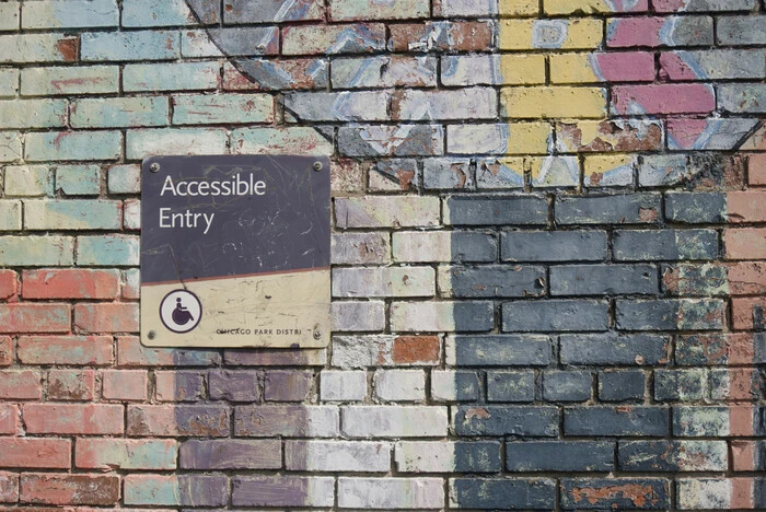 A colorful brick wall features a sign with "Accessible Entry" and a wheelchair symbol. The background is painted with various abstract shapes and colors, including blue, pink, yellow, and white, creating a vibrant, textured effect.