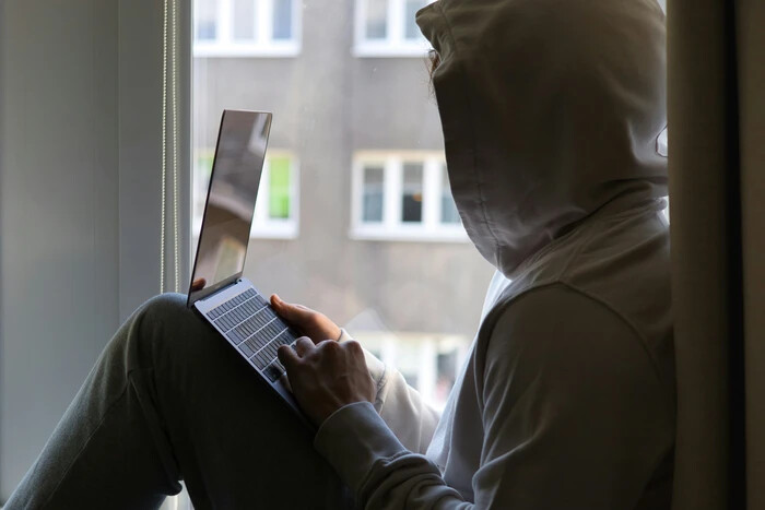 Person in a hoodie sits by a window, intently using a laptop. The background shows an out-of-focus exterior with building windows.