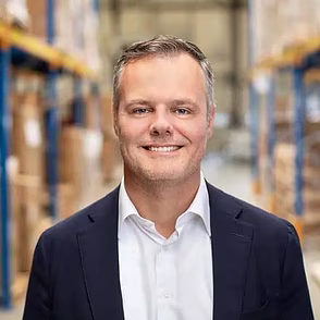 Man in donkerblauw jasje glimlacht in magazijn met stellingen op de achtergrond